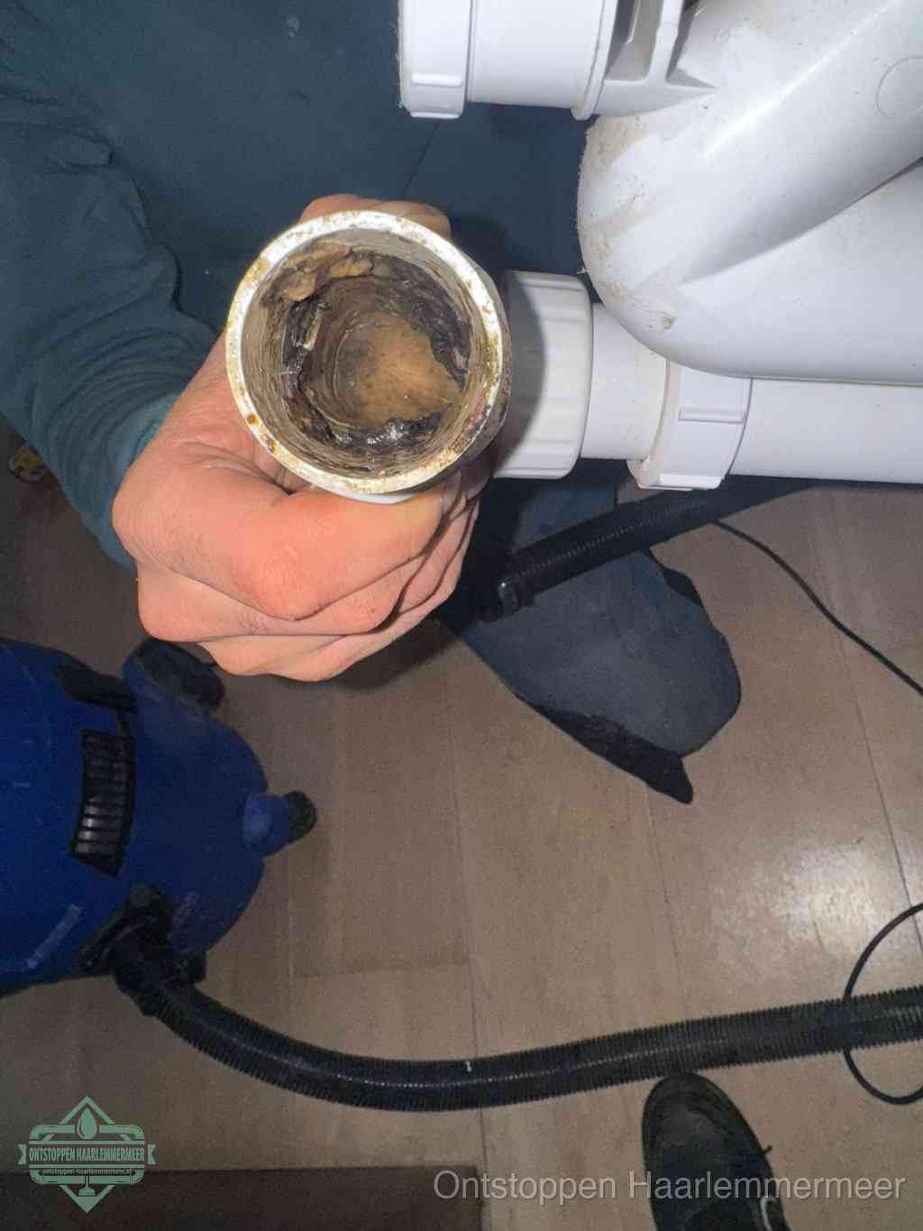 Een hand houdt een losgekoppeld PVC-afvoerbuisstuk vast dat van binnen volledig is dichtgeslibd met vet, vuil en aanslag, de meest voorkomende oorzaak van een verstopte afvoer in Haarlemmermeer
